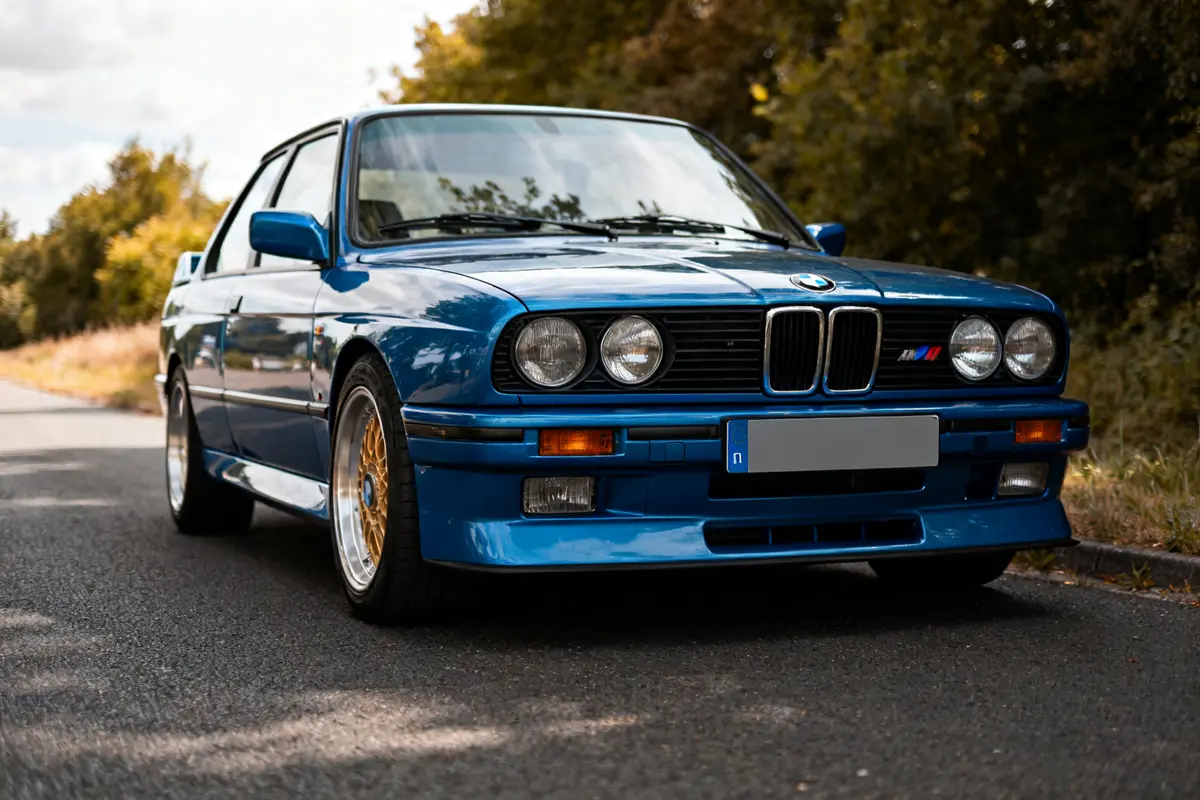 BMW ancienne bleue, modèle sportif, sur route entourée d'arbres verts en arrière-plan
