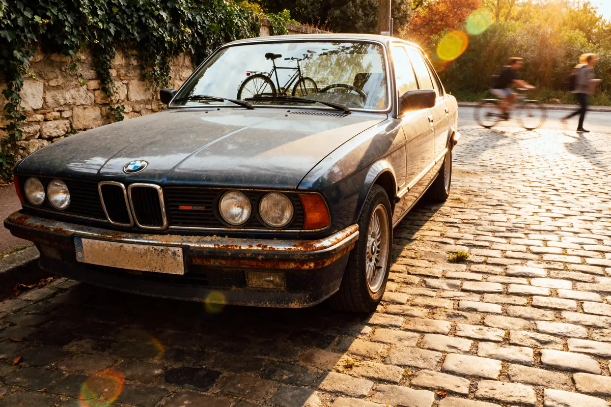 BMW e28 automatic stationnée sur une rue pavée ensoleillée avec cyclistes à l'arrière-plan