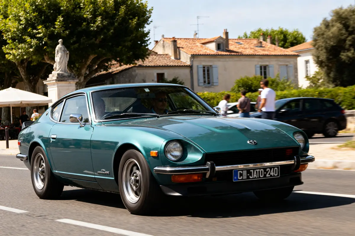 Voiture classique verte Datsun 240z automatic sur route au soleil devant bâtiments historiques