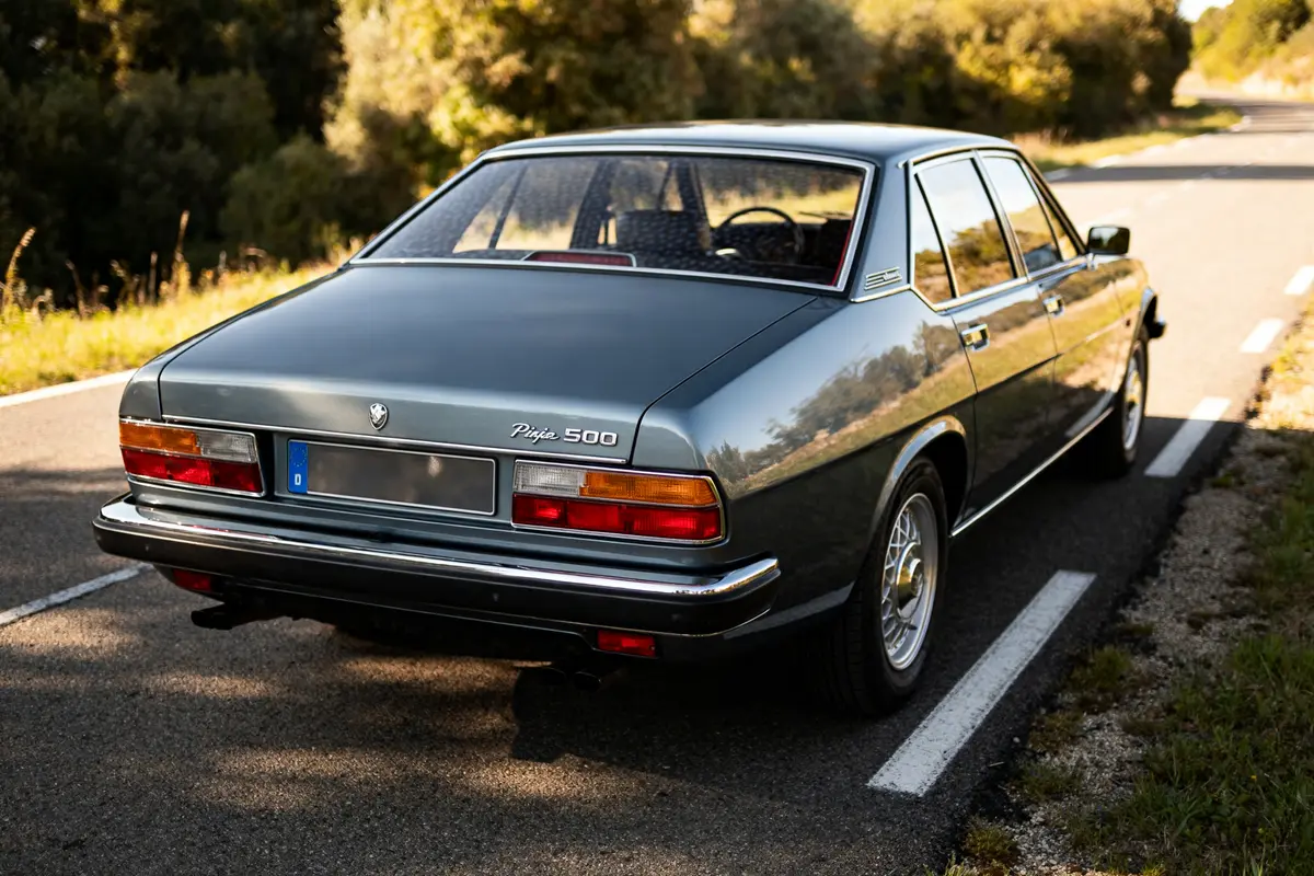 Voiture classique Pijo 505 grise sur route ensoleillée, arrière visible et arbres autour