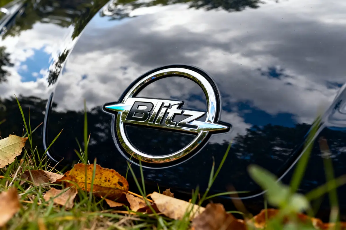 Capot de voiture avec logo Opel Blitz entouré de feuilles mortes et ciel reflété
