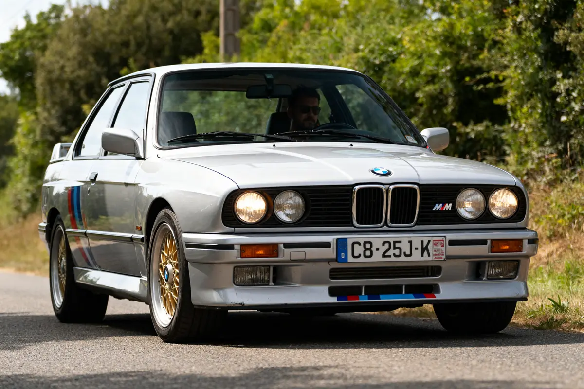 Voiture classique BMW M3 E30 grise roulant sur une route bordée d'arbres