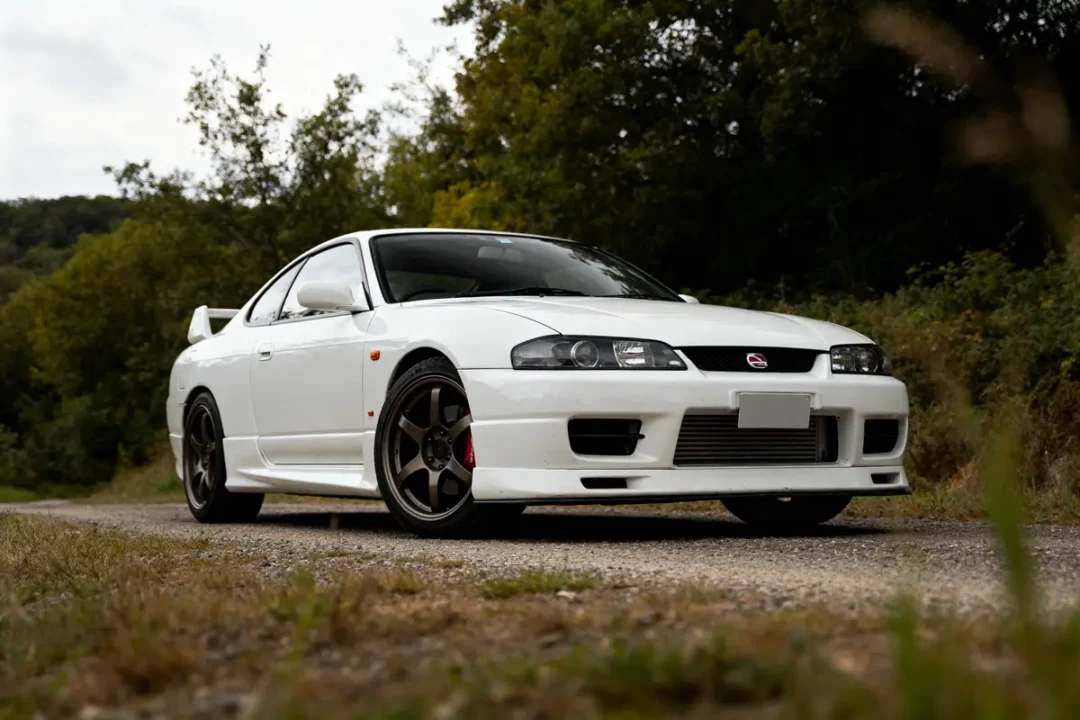 Voiture blanche sur route devant forêt, ressemblant à une Nissan Silvia S15 sportive.