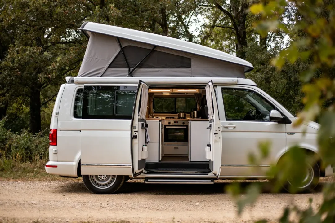 VW the California, van aménagé, toit relevé, portes arrière ouvertes en forêt verdoyante