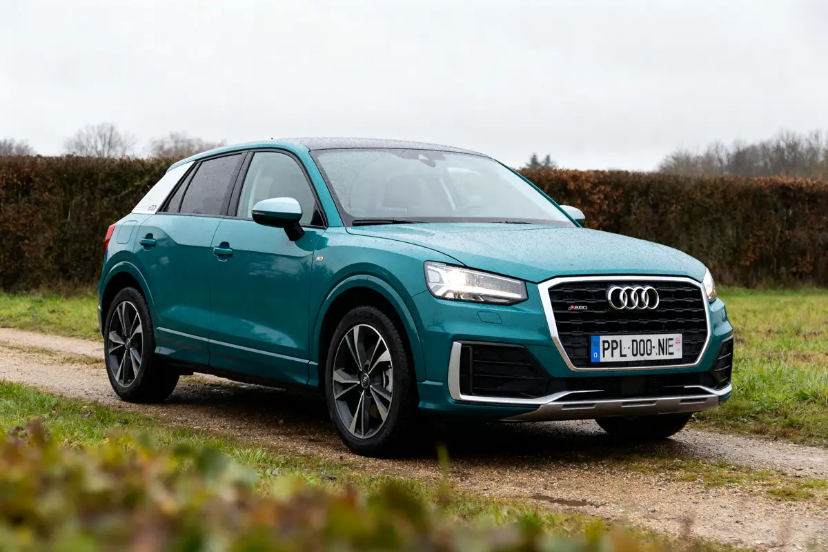 SUV Audi turquoise sur chemin de campagne, fond végétation et ciel nuageux.