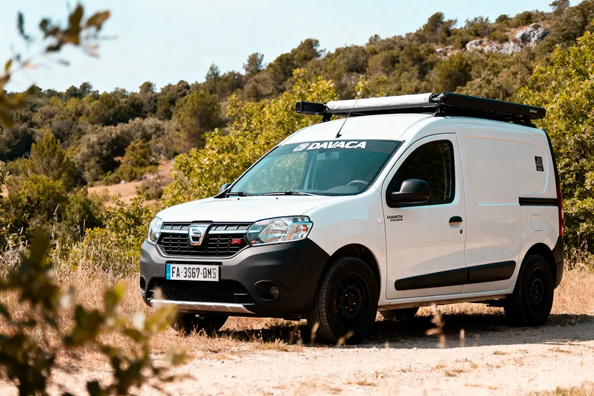 Un fourgon blanc, marque Dacia, stationné sur un chemin rural entouré de végétation verdoyante.