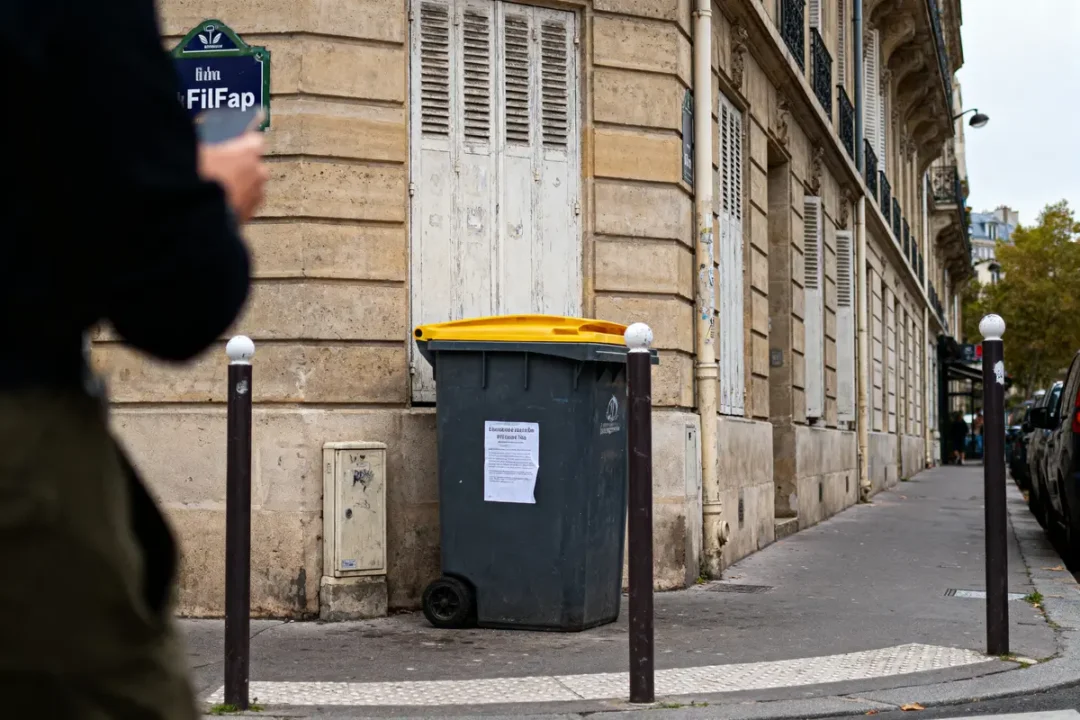 Homme flou avec smartphone près d'un conteneur à déchets sur le trottoir parisien, rue en arrière-plan.