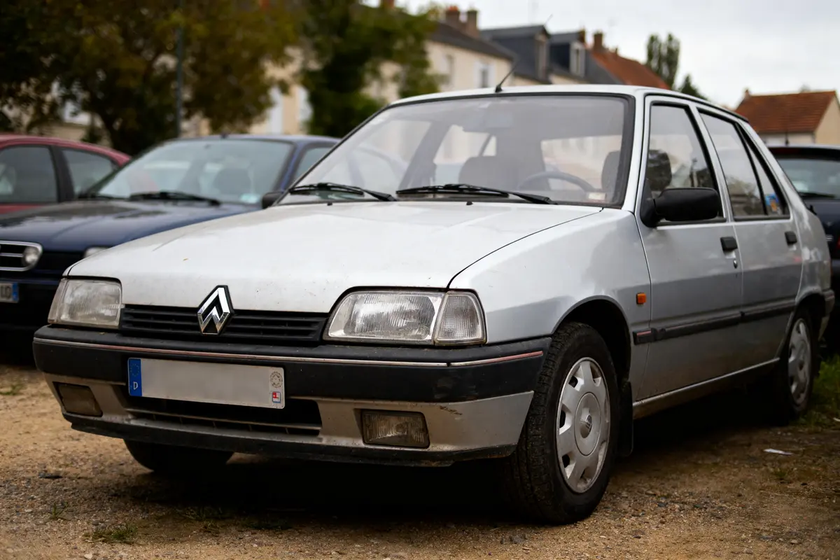 Voiture ancienne, grise, stationnée à l'extérieur, plaque floue. Arrière-plan avec arbres et maisons floues.