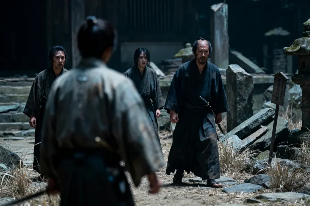 Quatre personnes en kimono sombre se font face dans un paysage de ruines et de pierres.