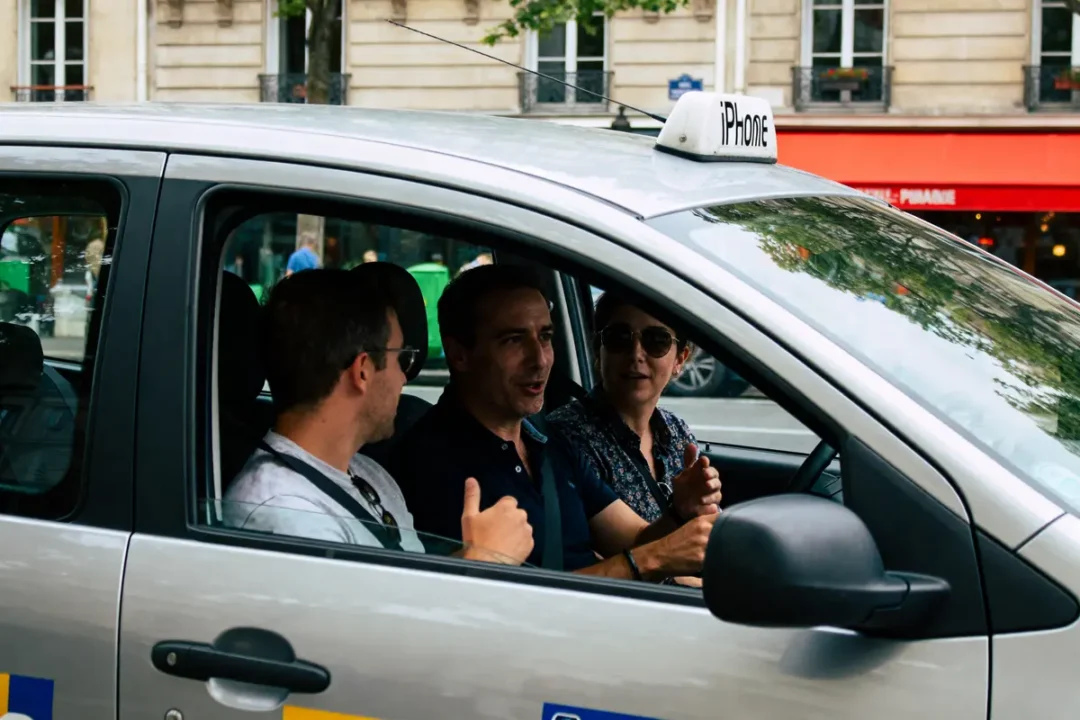 Trois personnes dans une voiture discutent en conduisant dans un cadre urbain. Panneau "iPhone" sur le toit.