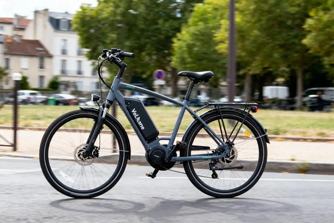 Vélo électrique gris stationné sur une route, avec arbres en arrière-plan urbain.